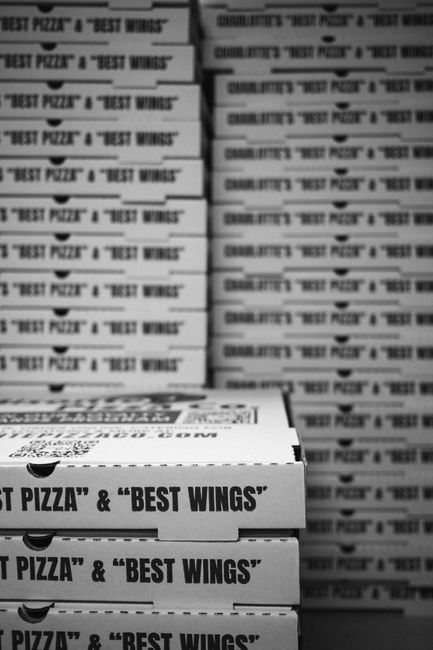 A stack of pizza boxes with the label 'Best Pizza' stacked horizontally on a surface, and more boxes in the background arranged vertically, creating a repeating pattern. The boxes are white with black
