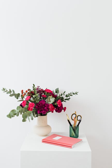 A floral arrangement featuring vibrant pink and deep magenta carnations, complemented by sprigs of greenery with broad, silvery leaves, placed in a simple, off-white ceramic vase on a white table. To 