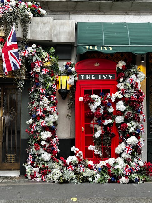 A vibrant floral display surrounding a classic red British telephone booth outside The Ivy restaurant, featuring white, red, and blue flowers arranged in a lush, cascading bouquet. The display is ador