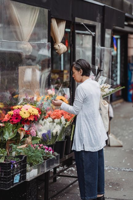 A woman standing outside Fitzrovia Flowers, a floral shop near Goodge Street station, examining a vibrant arrangement of fresh flowers. The display includes bright yellow, pink, and white daisies, alo