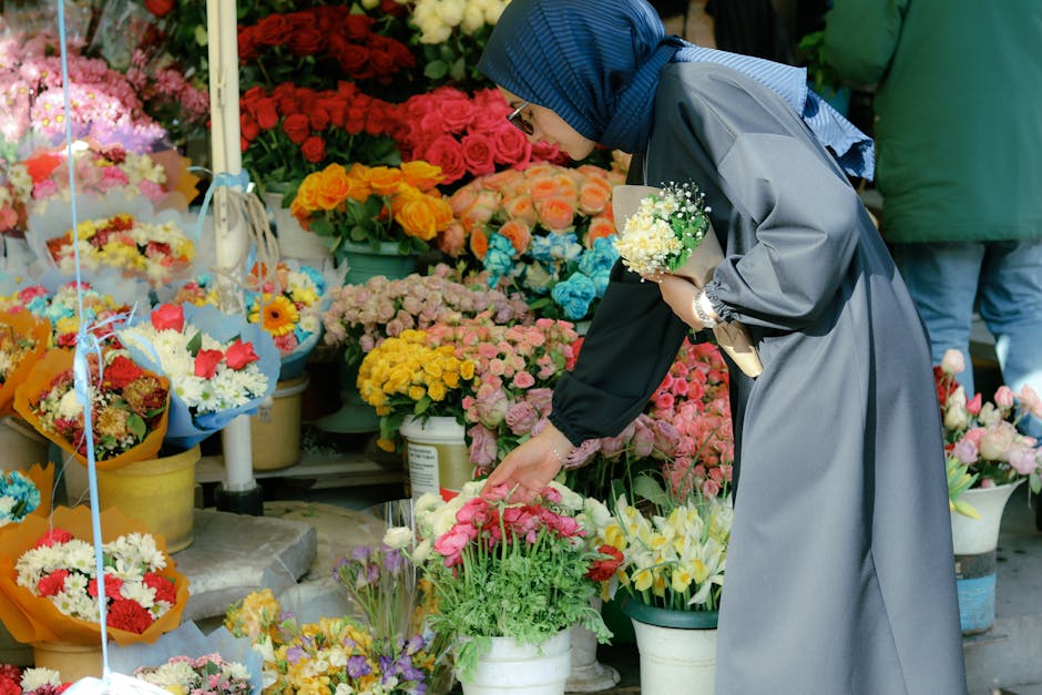 A woman wearing a blue headscarf, glasses, and a grey floral-patterned raincoat chooses a bouquet of pink and white flowers at a vibrant flower stall. The stall is filled with an array of fresh, color
